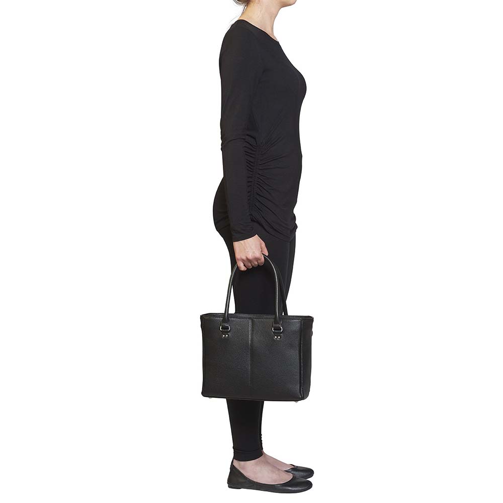 model from the side holding a Black timeless Traditional Elegance Concealed Carry Purse with pebble-textured cowhide leather. Features 11-ply steel cable reinforced, slash-resistant carry handles, designed for left or right-handed use. Spacious interior with center zippered divider, leather-trimmed pockets, and zippered pocket. Magnetic snaps close the open top. Includes metal feet for bottom protection. Khaki GTM logo lining for easy visibility. Includes holster compatibility.