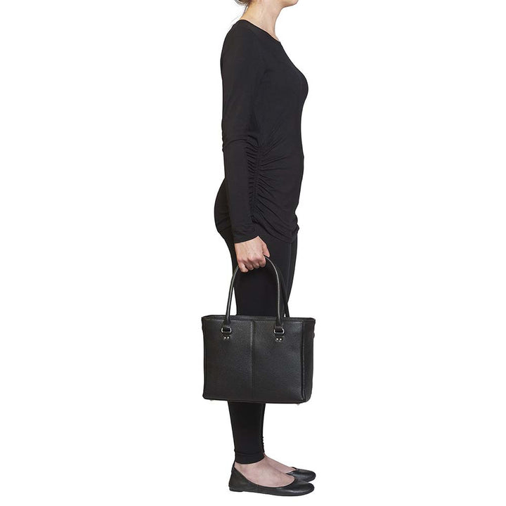 model from the side holding a Black timeless Traditional Elegance Concealed Carry Purse with pebble-textured cowhide leather. Features 11-ply steel cable reinforced, slash-resistant carry handles, designed for left or right-handed use. Spacious interior with center zippered divider, leather-trimmed pockets, and zippered pocket. Magnetic snaps close the open top. Includes metal feet for bottom protection. Khaki GTM logo lining for easy visibility. Includes holster compatibility.