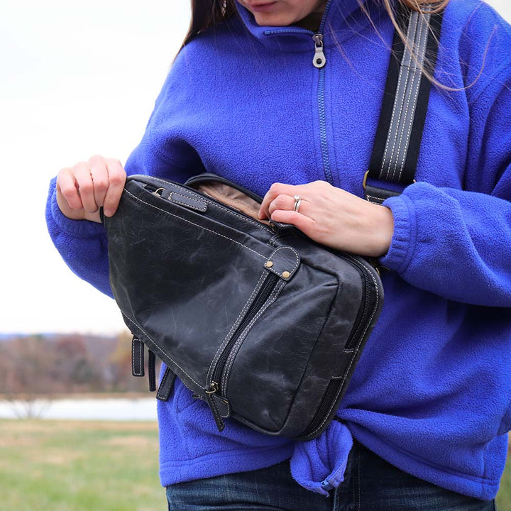 woman opening and looking inside of a Gray sling backpack concealed carry bag with RFID protection, ambidextrous draw, and padded CCW compartment; lightweight crossbody made for quick access, featuring slash-resistant strap, fits Glock 19, Sig P365, M&P 9mm, and ideal for everyday or EDC use.