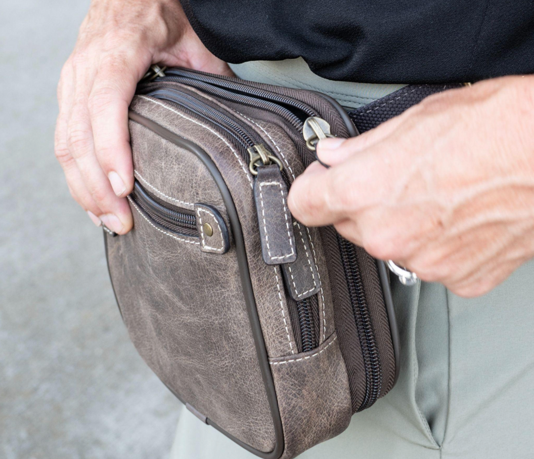 Close-up of hands holding a rugged brown leather crossbody bag with heavy-duty zippers.