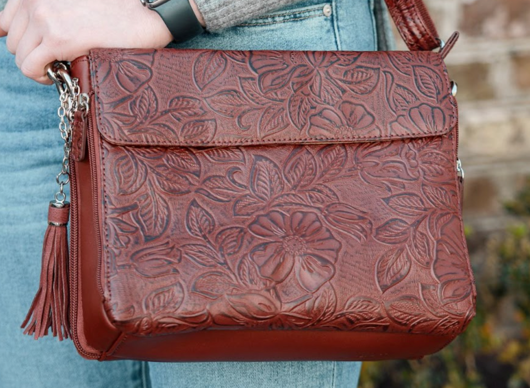Close-up of a brown embossed leather purse with a floral design, worn over a light top.