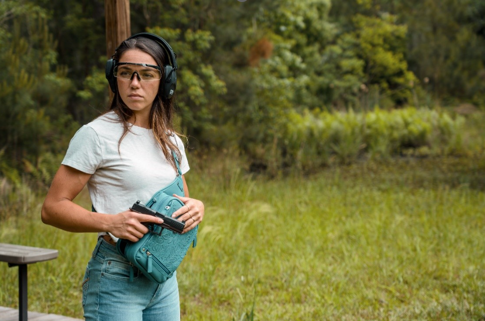 Woman at an outdoor shooting range holding a teal concealed carry purse, wearing protective glasses and ear protection.