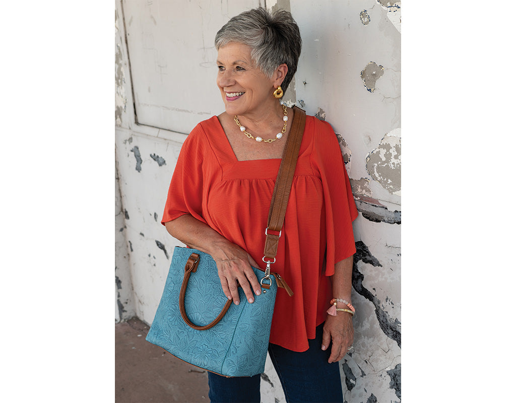 Smiling woman in a red top and jeans standing against a white wall, holding a blue concealed carry handbag.