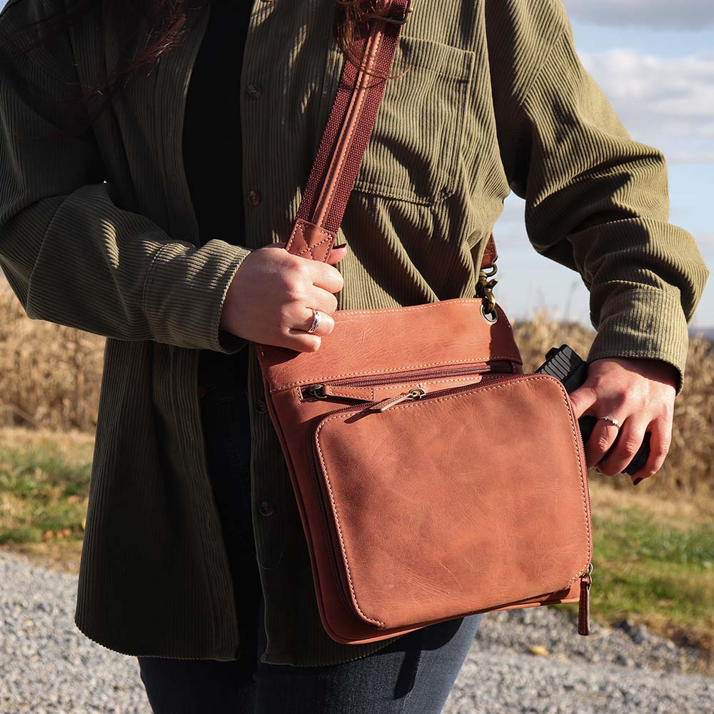 close up of model pulling weapon out of Vintage distressed buffalo leather concealed carry crossbody purse with adjustable slash-resistant strap, left and right-handed access, and a secure gun compartment designed for fast draw. Includes holster and fits compact handguns like S&W Shield, Ruger LCP, and Glock 42.