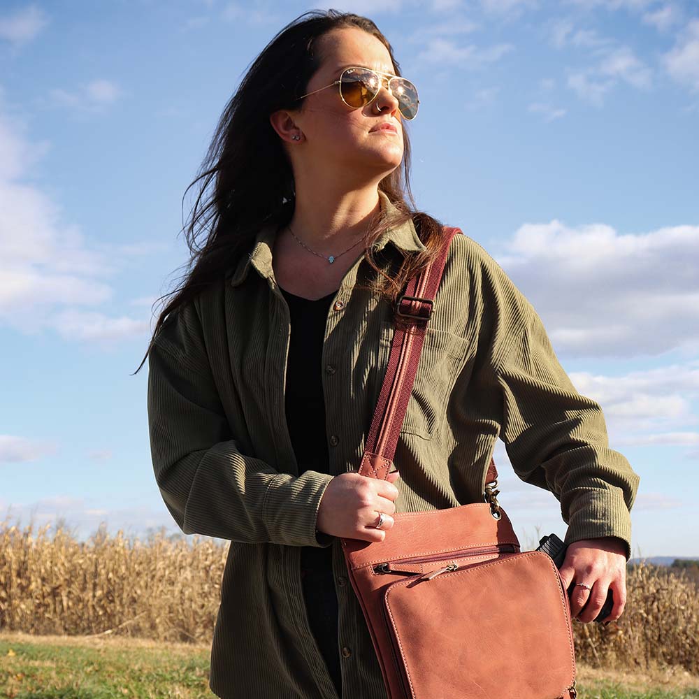 model walking in front of field with Vintage distressed buffalo leather concealed carry crossbody purse with adjustable slash-resistant strap, left and right-handed access, and a secure gun compartment designed for fast draw. Includes holster and fits compact handguns like S&W Shield, Ruger LCP, and Glock 42.