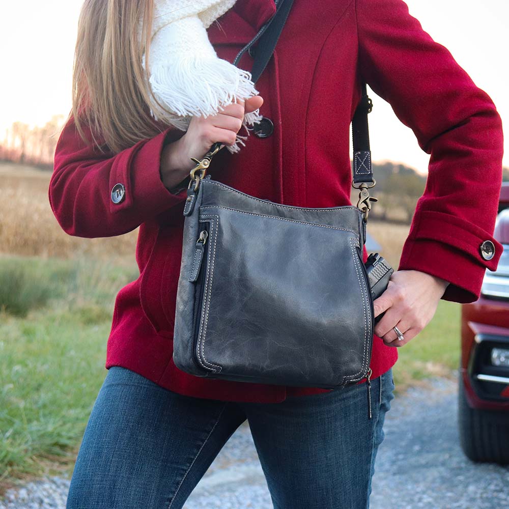 woman grabbing a gun out of a Gray distressed leather crossbody concealed carry bag with RFID-blocking card storage, vertical top-draw gun access, supports left/right-handed draw, secure fit for compact handguns, anti-slash strap included