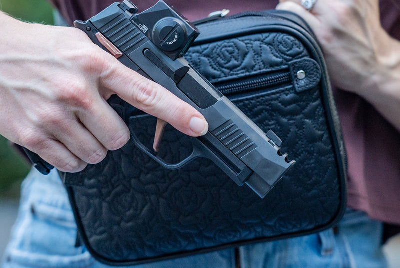 A close-up lifestyle shot of a person holding a black semi-automatic pistol in one hand while holding a black quilted zippered case with the other hand. The person is wearing a maroon t-shirt and blue jeans, with a blurred outdoor background.