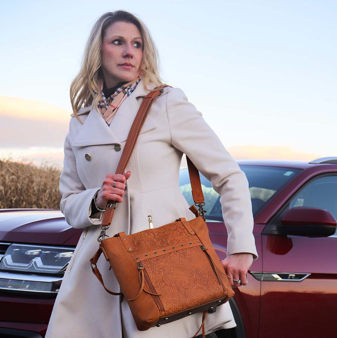 woman pulling out a concealed pistol out of a Tan concealed carry BOHO tote purse with top-access CCW compartment, symbolic butterfly and flower debossed leather, slash-proof straps, RFID-blocking zip pocket, and room for compact pistols like CZ P10, M&P Shield, and S&W EZ—each bag uniquely aged from natural tanning.