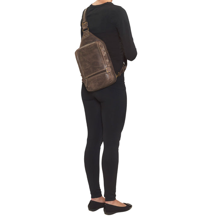 model from behind with Distressed brown buffalo leather sling concealed carry backpack with tactical organization, RFID zip pocket, padded holster compartment for Glock 19, Sig P320, and HK VP9, slash-proof strap, and swing-front crossbody design with 3 exterior zip pockets and morale patch zone